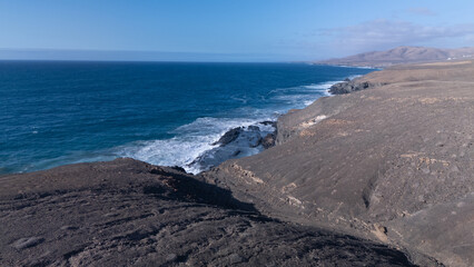 Rocky coastline with powerful waves crashing against rugged cliffs under a clear sky, showcasing...