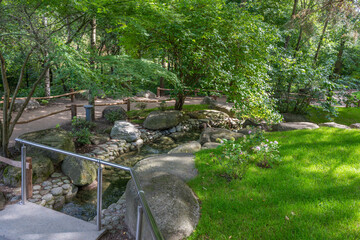 A small stream flows between rocks in a landscaped garden. Green grass and trees create a calming atmosphere.