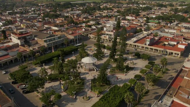 Upward aerial from central kiosko revwals symmetrical park layout, palm-lined streets, and surrounding town buildings in Tuxpan, Jalisco