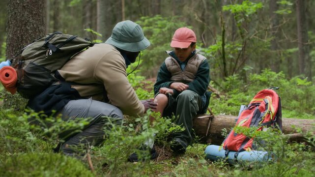Caring African American father helping his little son by putting bandage on his scraped knee while hiking in woods, showing support and care on their outdoor adventure