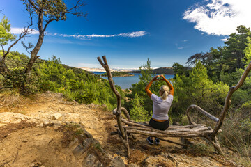 A woman sits on a bench in the middle of a forest, with a panoramic view of Strucic Beach on the...