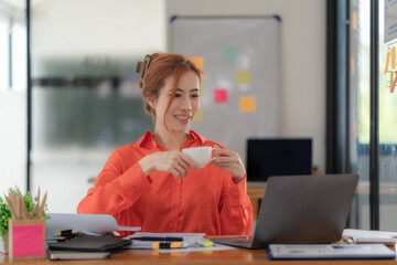 Smiling woman drinking coffee while working on a laptop in a modern home office.