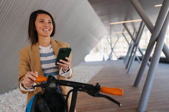 Smiling asian woman outdoors with smartphone and electric scooter