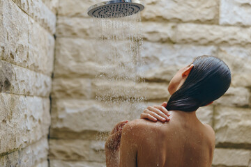 woman taking shower under rainfall showerhead in modern bathroom with stone walls, enjoying...