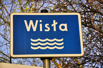 Niebieski znak drogowy z nazwą rzeki „Wisła” i symbolem fali wodnej na tle gałęzi drzew i nieba. Idealny do przedstawiania koncepcji nawigacji, punktów orientacyjnych i oznakowania kierunkowego. © Pol Badger