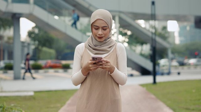 Portrait of hijab wearing woman navigates via smartphone walks in urban park. Happy Malaysian female office employee reads work chat on mobile phone strolling on street. City life