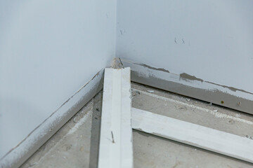 Corner of room during renovation showing partially removed baseboards with nails exposed, scattered...