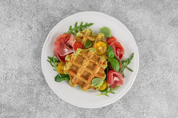 Spinach waffles with tomatoes, arugula and prosciutto on a white plate with a cup of coffee....