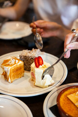 Hands sharing assorted mini cakes on a white plate, including lamington, caramel square, and strawberry shortcake slice, cozy dessert moment at table