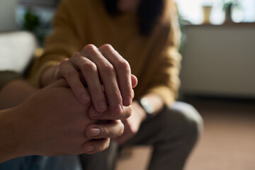 Caucasian young adult woman gently holding hands with Caucasian young adult man offering comfort and support, both seated indoors with focus on hands and blurred background