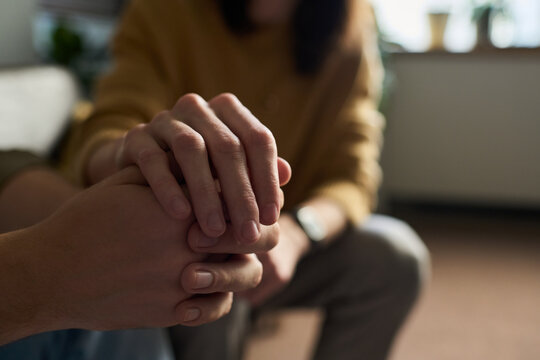 Caucasian young adult woman gently holding hands with Caucasian young adult man offering comfort and support, both seated indoors with focus on hands and blurred background