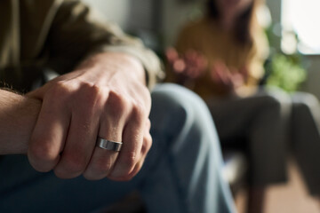 Caucasian young adult man wearing wedding ring sitting with hand in focus, while blurred young...