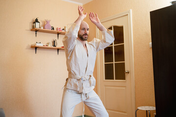 Man in traditional martial arts uniform practicing an exercise with raised hands indoors,...