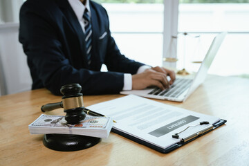 A professional lawyer in a suit works on a laptop with legal documents, gavel, money, and scales of justice on desk.