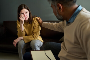 Caucasian middle aged woman sitting on couch wiping tears while Indian middle aged man comforting...