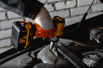 A mechanic wearing protective gloves holds a cordless wrench next to a car engine. Concept with...