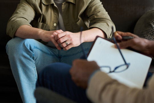 Caucasian young adult man sitting with hands clasped during counseling session, middle aged man holding notebook and eyeglasses conducting professional consultation