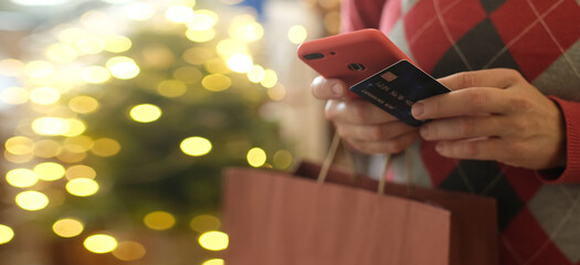 Online Christmas shopping. Widescreen banner, copy space. Woman holding shopping bag making payment using bank card and mobile phone standing in front of bokeh lights.