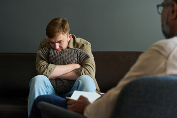 Caucasian young adult man sitting on couch hugging pillow, looking down with worried expression...