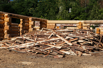 Wood scraps (bark, offcuts) piled before a partially built log house. Shows materials & byproducts...