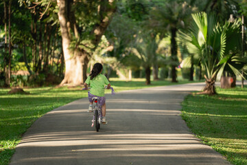 Fototapeta premium Young girl riding bicycle on a serene pathway in a lush green park surrounded by trees and sunlight filtering through the leaves creating a tranquil scene