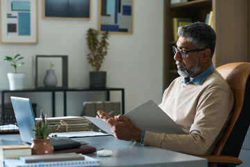 Middle aged Indian man sitting at desk reviewing documents while working on laptop in modern...
