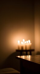 A group of lit candles on a small wooden table, casting a warm glow in a dark room, with a larger table in the foreground.