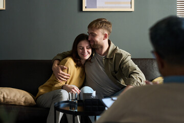 Caucasian young adult man embracing Caucasian young adult woman on couch, both smiling with closed...