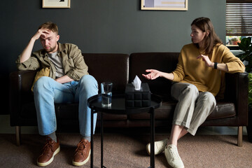 Caucasian young adult man sitting on sofa holding head looking distressed while Caucasian young...