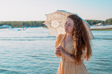 A young woman in a yellow dress under an umbrella on the beach. Portrait of a woman 30-35 years old. A sunny summer day. Romantic photo of a walk under a white cloth umbrella.
