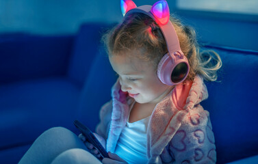 A young girl wearing glowing cat-ear headphones and a cozy pink jacket sits on a couch, happily...