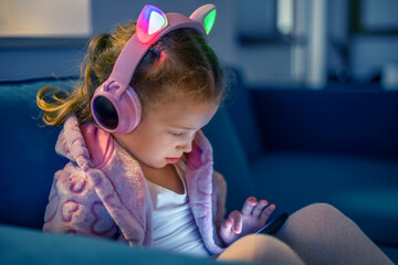 A young girl wearing glowing cat-ear headphones sits indoors on a sofa, focused on a digital device...