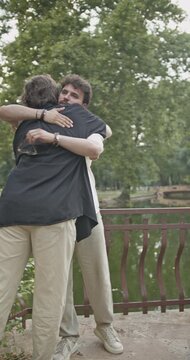 Two young men, good friends, meet and share a warm hug before engaging in a friendly conversation in a lush green park near a serene lake. They appear happy and relaxed.