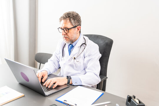 Mature male doctor with stetoscope working while looking pc in medical consultation in doctor’s office – senior doctor on laptop computer online for hospital schedule  - Powered by Adobe
