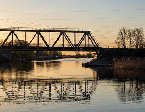 Silhouettes of cyclists crossing a steel truss bridge over a calm river during a golden spring sunset with water reflections.