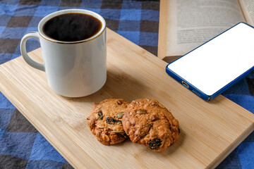 Relaxing top view of black coffee in a white ceramic mug with homemade cookies on a wooden cutting board, placed beside a smartphone and an open book. Perfect for lifestyle, cafe, and digital design