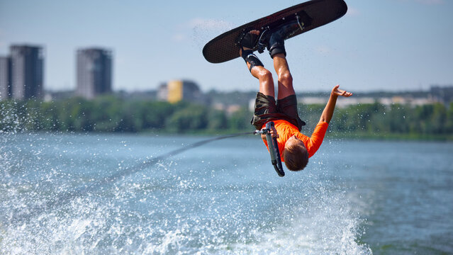 Boy wakeboarder upside down flip stunt on river in bright orange shirt. Concept of adrenaline, extreme water sports, youth courage, balance, and emotional thrill. - Powered by Adobe
