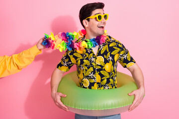 Cheerful young man holding an inner tube and wearing tropical print shirt with decorative lei on...