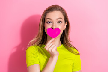 Cheerful young woman holding a pink heart while smiling against a vibrant pink background wearing a...