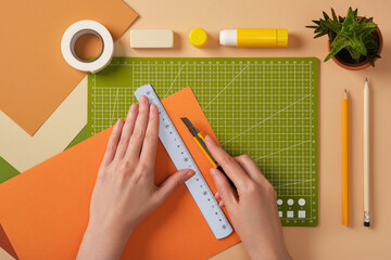 Hands cutting orange paper with a ruler and knife on a green mat. The process of creating art from...