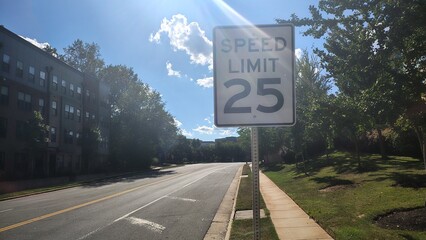 A 25 mph speed limit sign on a road between a park and an apartment building in the United States on a sunny day, American traffic rules and speed regulations, road signs on American urban highways