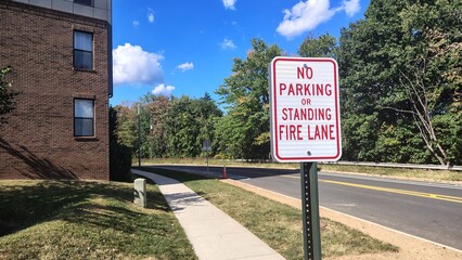 A road sign forbidding parking and stopping in the fire lane is located to the right of the brick building in front of the trees along the road.
