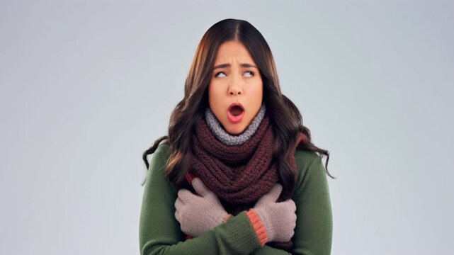 A woman wearing winter clothing including a scarf and gloves pretending to feel cold while breathing out visible air and shivering gently against a pure white background in a high key studio setup