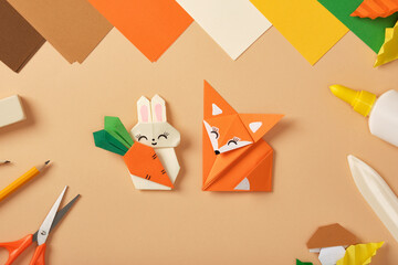 Paper rabbit with carrot and fox origami figures on desk with colored sheets, scissors, pencil,...