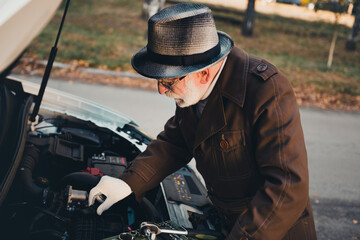 Elderly man inspecting car engine during a sunny autumn day outdoors in a park surrounded by...