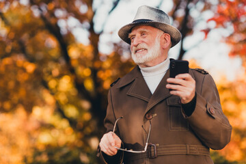 Senior man enjoying an autumn walk in the park while using a smartphone and holding his glasses in the golden sunlight