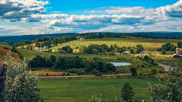 Preston County, West Virginia, USA | Rolling farmland dotted with hay bales stretches toward the horizon, where wind turbines trace the ridgeline beneath a sky layered with late-summer clouds.