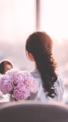 Young woman with long dark hair is sitting by a window, holding a bouquet of pink peonies, enjoying a serene moment of reflection and tranquility in a softly lit environment