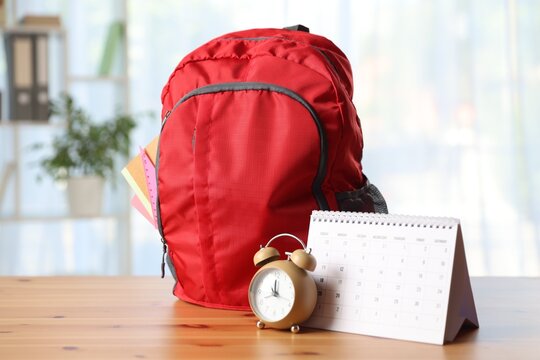 Red backpack with different stationery, calendar and alarm clock on desk in classroom - Powered by Adobe