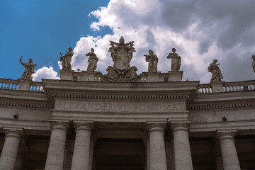 Architectural view of a building with statues, columns, a crest, and an inscription on a cloudy day. The sky is a mix of blue and grey.
